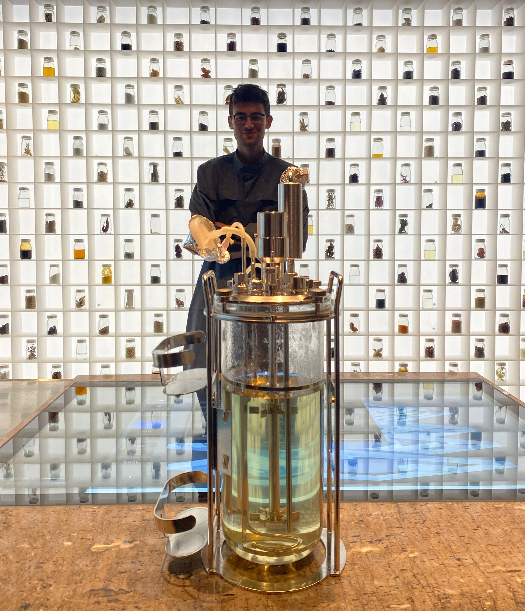 Hill-Maini in the test kitchen of Alchemist in 2021. The “Taste Wall” showcases different ingredients and experiments conducted by chefs at the Copenhagen restaurant over the years. He is standing behind a bioreactor that he and others experimented with to grow fungi.