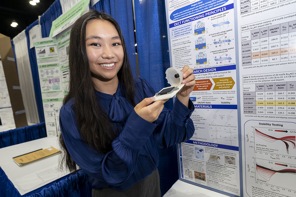 Grace Sun stands next to her project board at Regeneron ISEF 2024. She is holding a bioelectronic device that might one day help diagnose or treat health issues. 