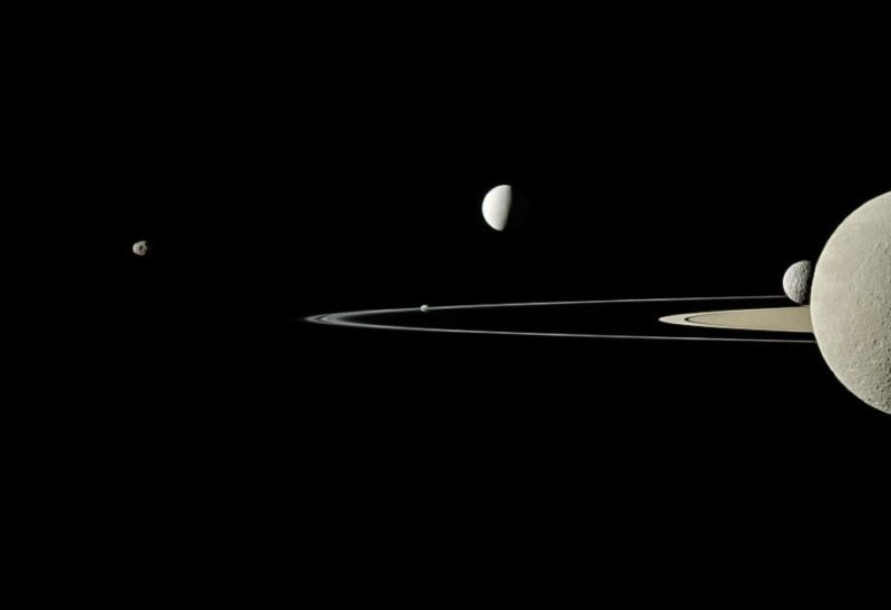 Saturn, the reigning champion of the solar system in terms of moons, now boasts 128 new additions! This image, captured by the Cassini spacecraft, features five of Saturn’s larger moons. Take a closer look—the cratered celestial body on the right isn’t Saturn but its moon, Rhea, positioned in front of the planet with some of Saturn’s rings visible in the background.
