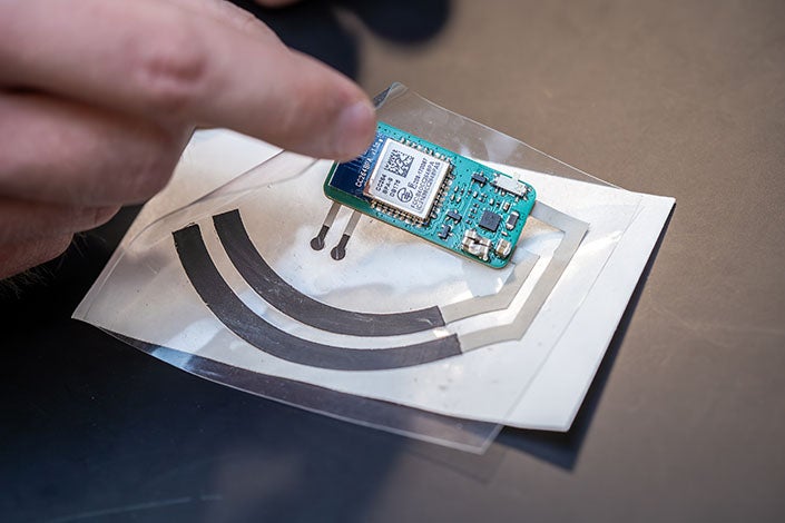 The sticker includes a biofuel cell (shown as black arches) beneath a porous hydrogel pad that gathers sweat from the fingertip, along with a printed circuit board and a vitamin C sensor (small black circles connected to the board).