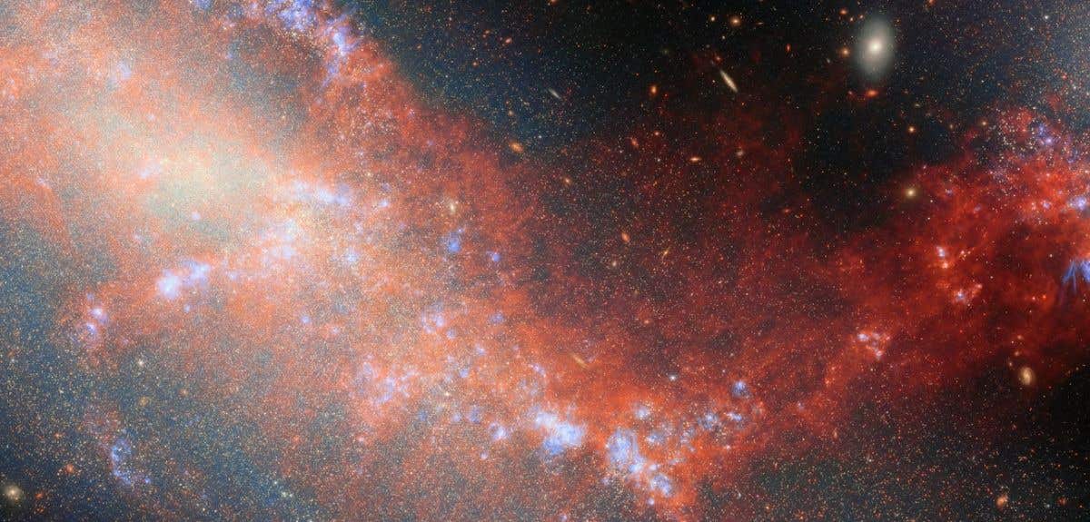 Image captured by the James Webb Space Telescope, showing two dwarf galaxies (NGC 4490 and NGC 4485, depicted as glowing hues) connected by a wispy bridge of gas (seen as a red stream). There are regions of bright blue ionised gas visible in concentrated areas of the red stream.