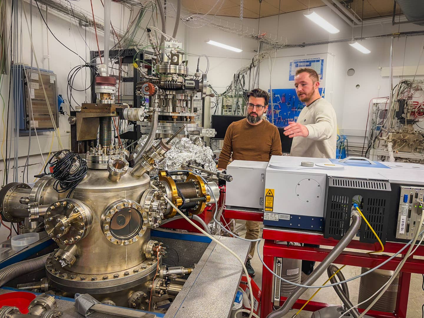 In the background, Associate Professor Sergio Ioppolo (left) and Postdoc Alfred Thomas Hopkinson (right) discussing experimental plans. In the foreground, two ultra-high vacuum chambers used to investigate reactions under interstellar medium conditions.
