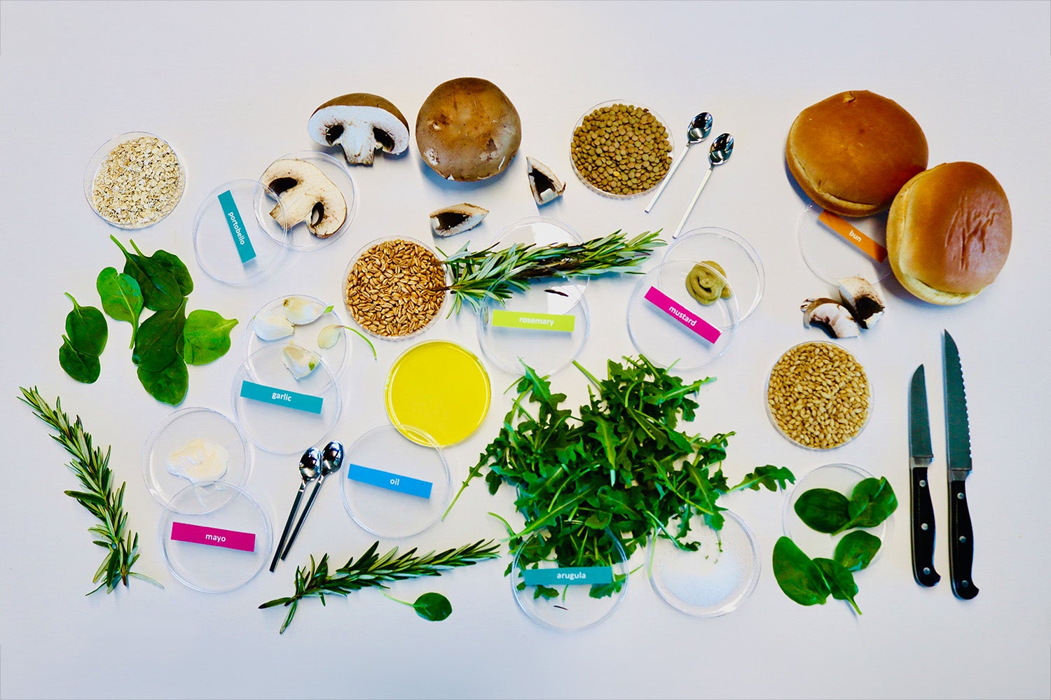 A selection of burger ingredients laid out on a table, including mushrooms, arugula, rosemary, and grains. 