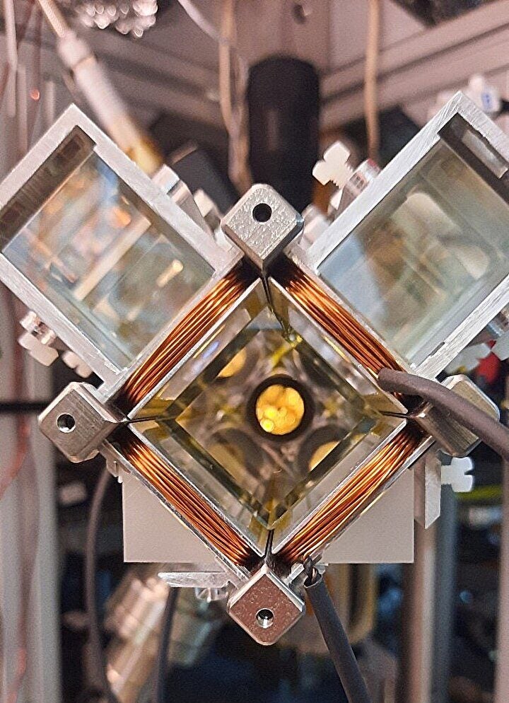 The 2D MOT apparatus which feeds the science chamber with cold atoms. At the centre is a glass cell held under vacuum between coils producing magnetic fields. The atoms go into the science chamber through the tiny hole that may be seen in the centre of the yellow (reflecting) surface