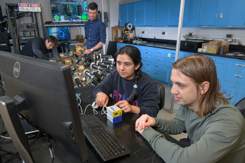 Doctoral students Masooma Sarfraz and Miles Garcia (center and right) examine data from CosmicWatch in the lab, while senior Collin Owens and Axani work on part of a future experiment that will incorporate the invention.